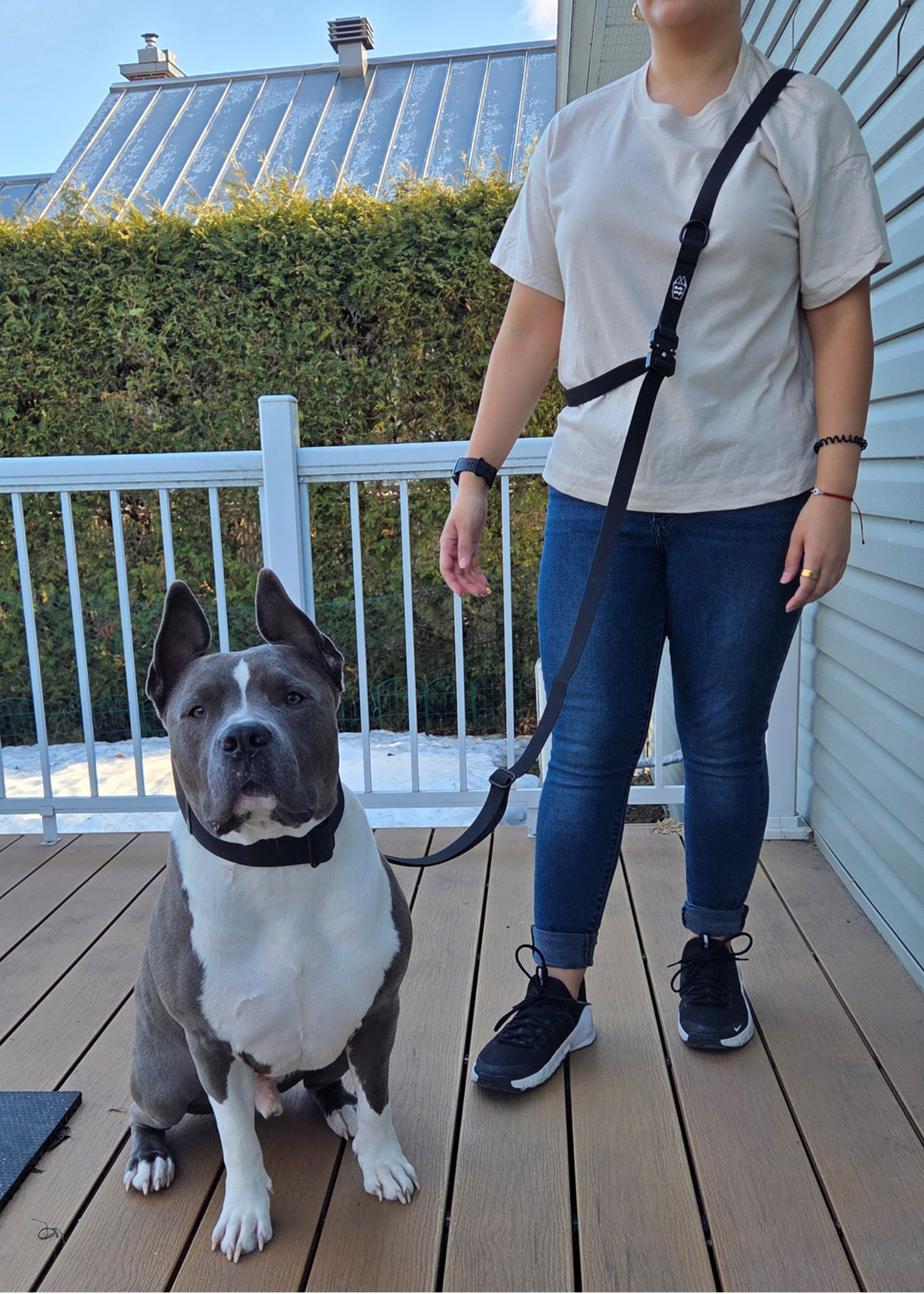 Person standing on a wooden deck with a dog on a leash, wearing a white shirt and blue jeans.