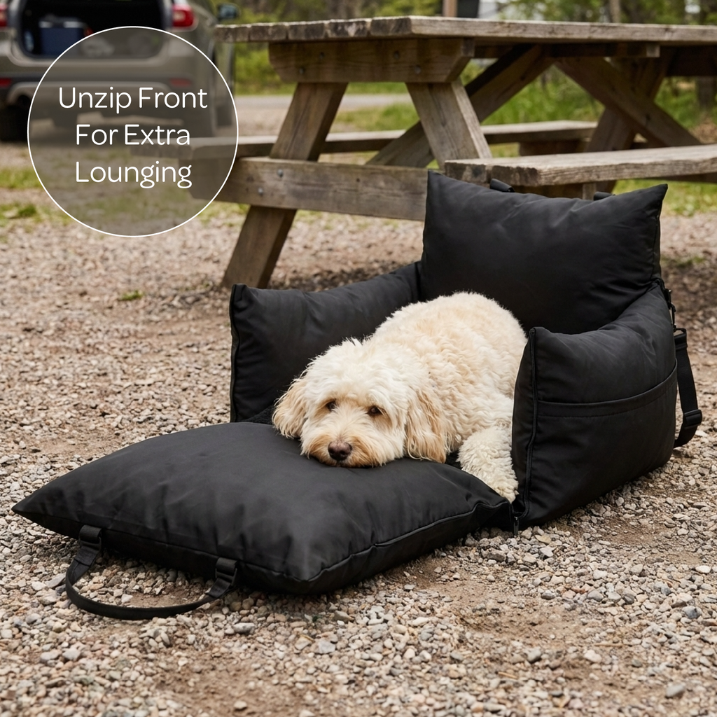 Dog lying on a black pet bed with an open front section, surrounded by a picnic table and gravel ground.