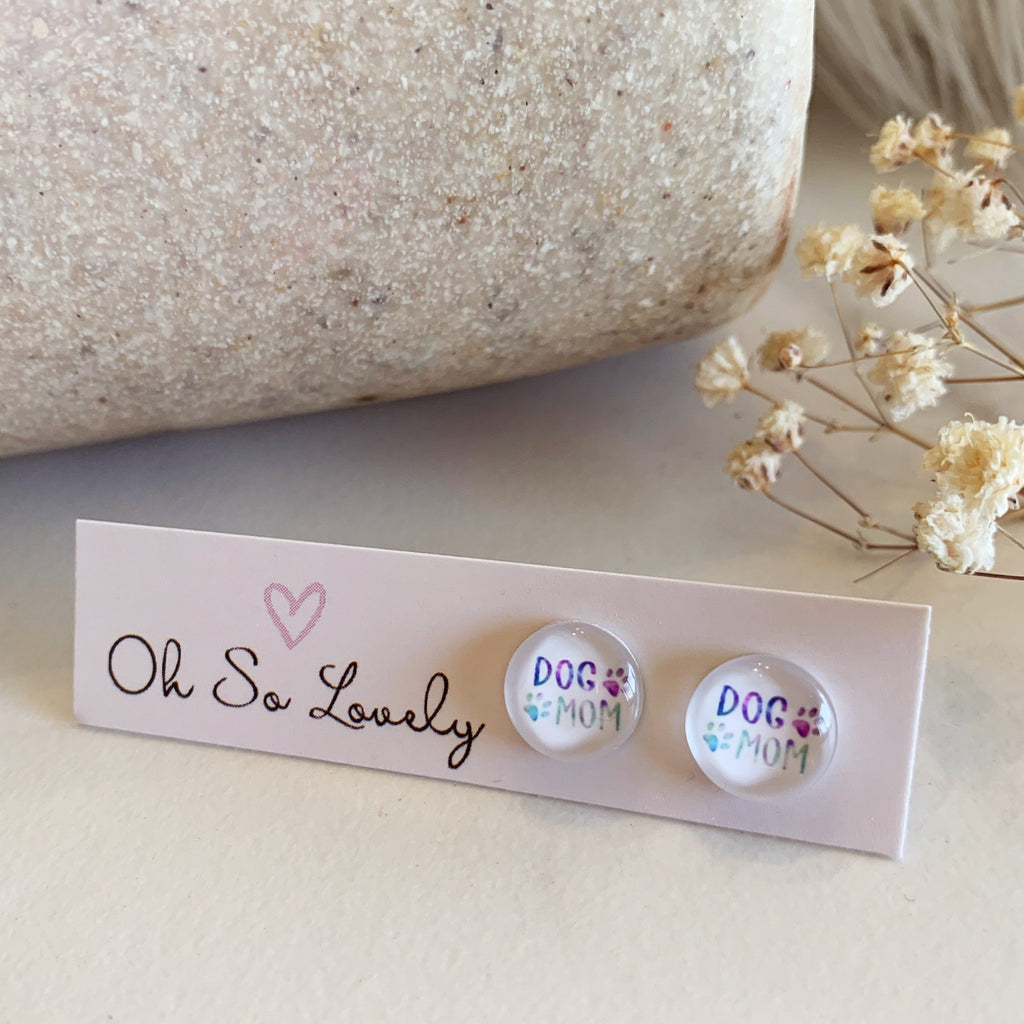 Two round earrings with 'DOG MOM' text on a card labeled 'Oh So Lovely' with dried flowers in the background.