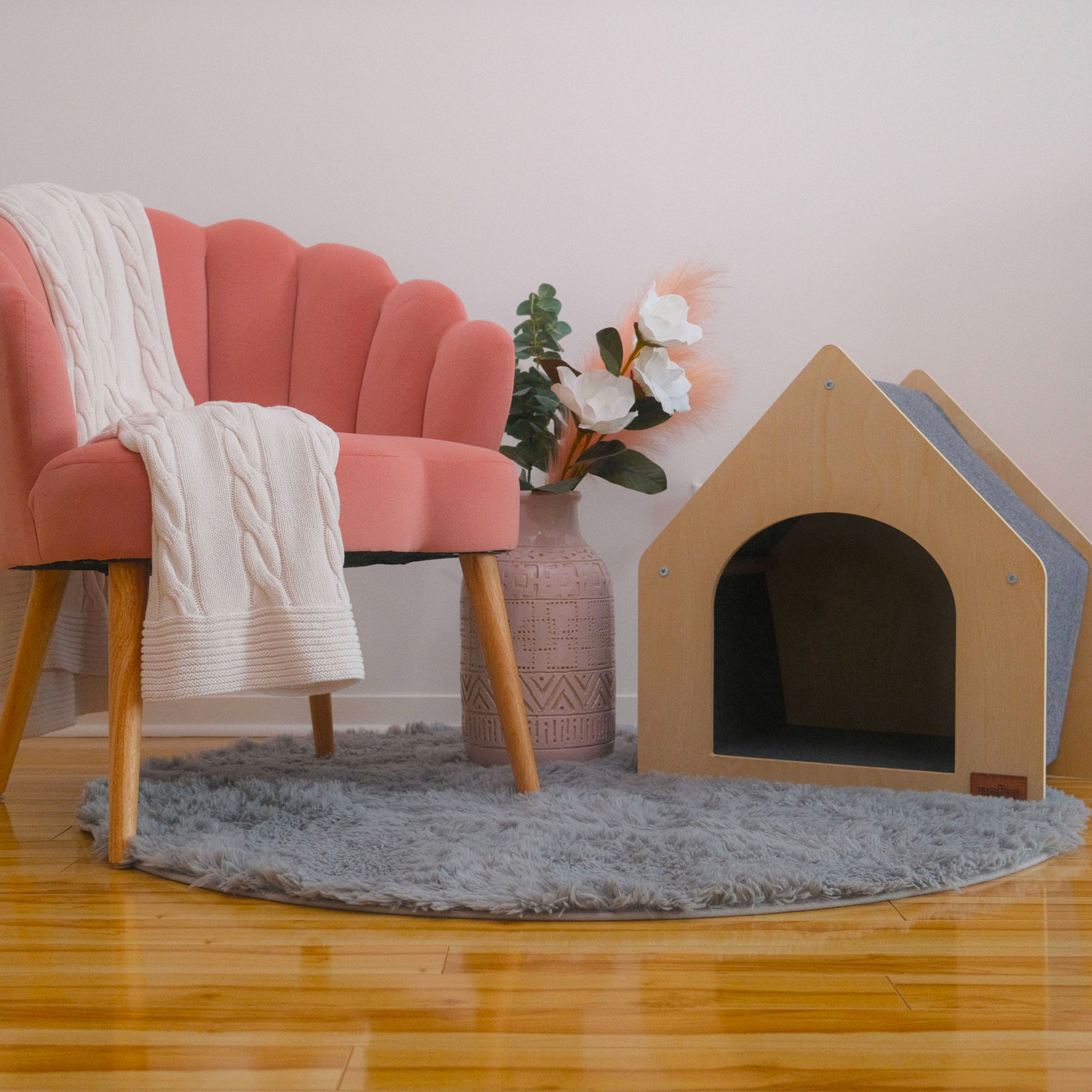 Pink chair with a white blanket, a vase with flowers, and a wooden pet house on a gray rug.