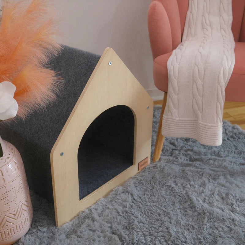 Wooden pet house on a gray carpet with a pink chair and decorative items in the background
