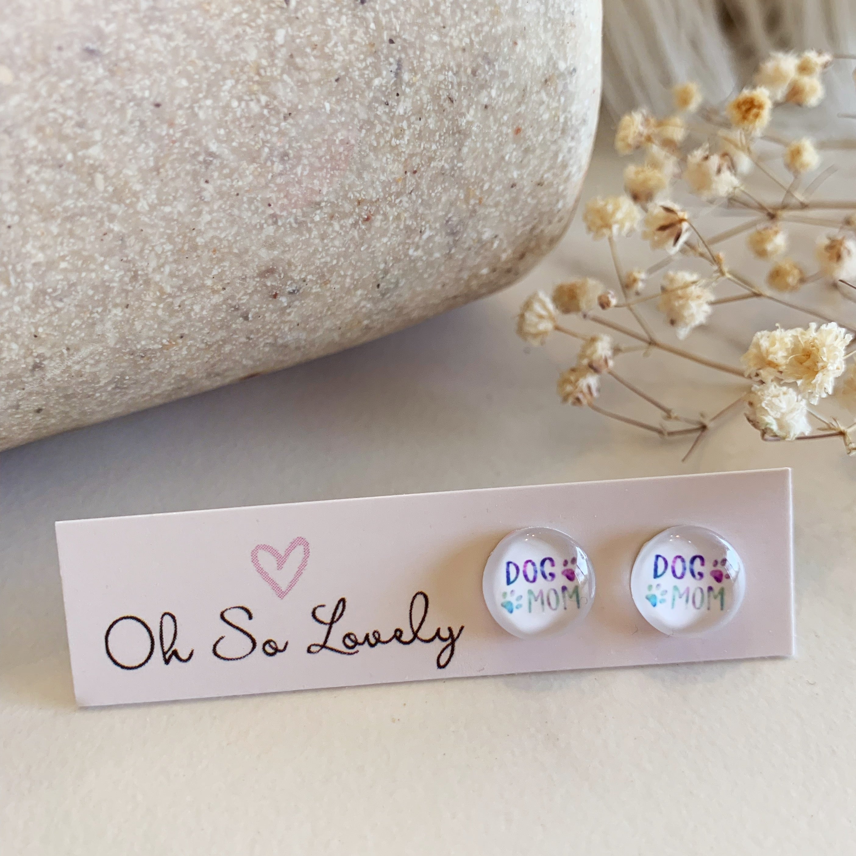Pair of earrings with 'DOG MOM' text on a 'Oh So Lovely' card with flowers in the background.