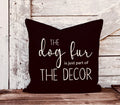Pillow with white text 'The dog fur is just part of the decor' on a wooden surface.