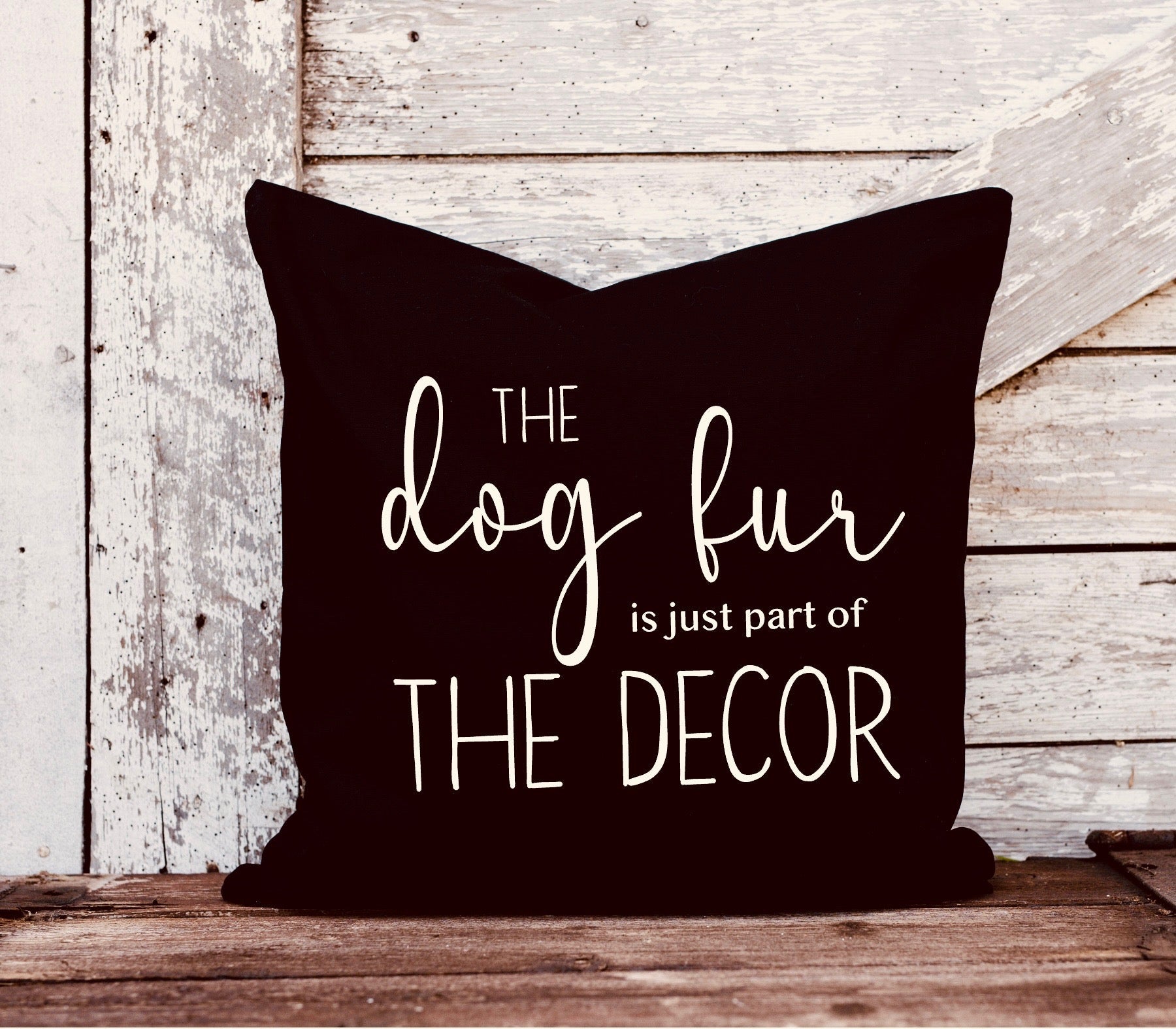 Pillow with white text 'The dog fur is just part of the decor' on a wooden surface.