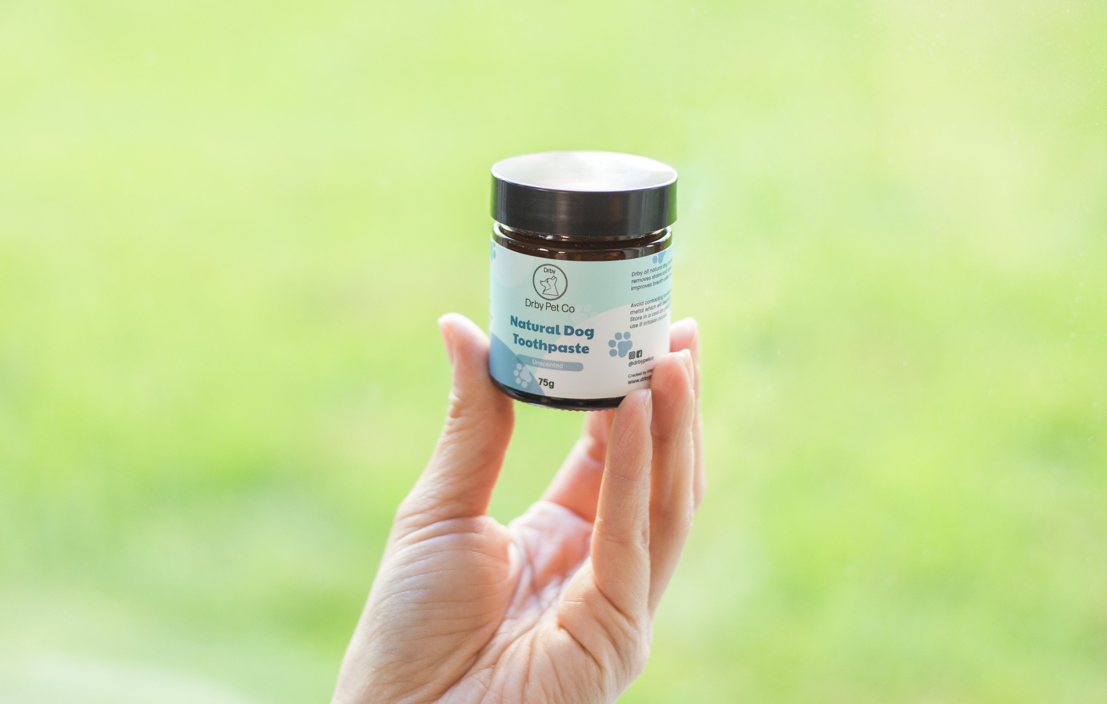 Hand holding a jar of 'Natural Dog Toothpaste' with a green blurred background