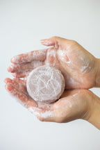 Hand holding a round, white, textured soap against a light background