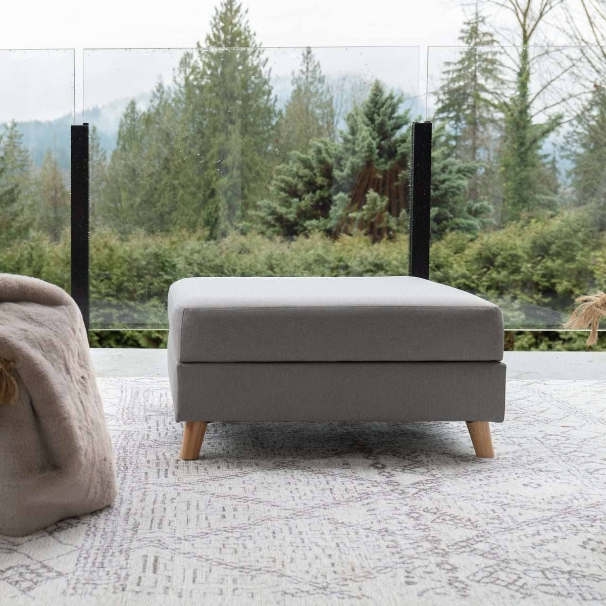Gray ottoman with wooden legs on a textured surface with a forest view in the background