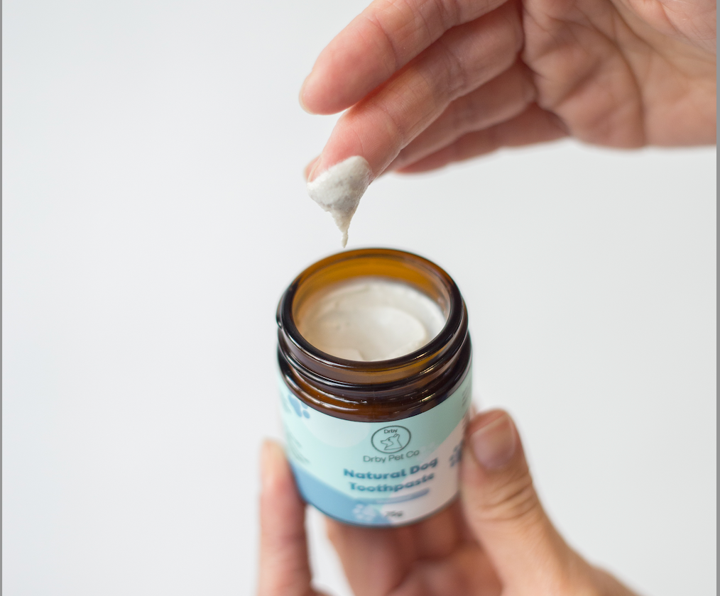 Hand holding a small jar of natural dog toothpaste with a person applying a white paste to a finger.