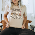 Person wearing a beige t-shirt with 'I Just Wanna Be a Stay at Home Dog Mom' text and graphics.