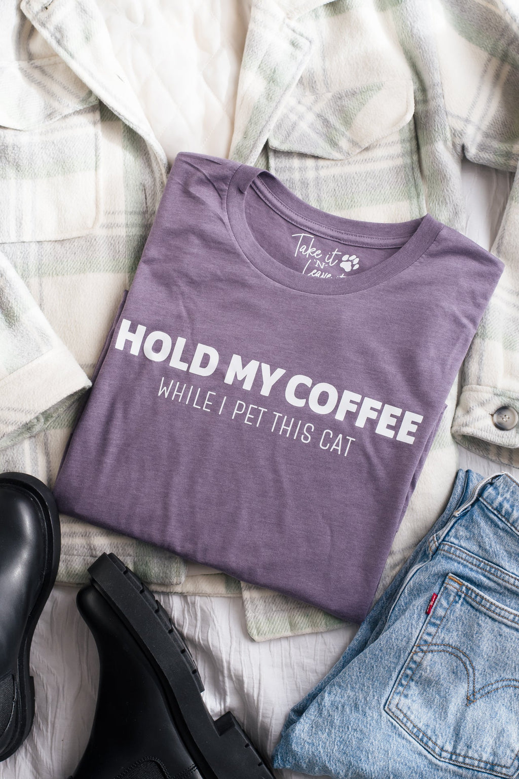 Purple t-shirt with text 'Hold my coffee while I pet this cat' on a checkered fabric background.