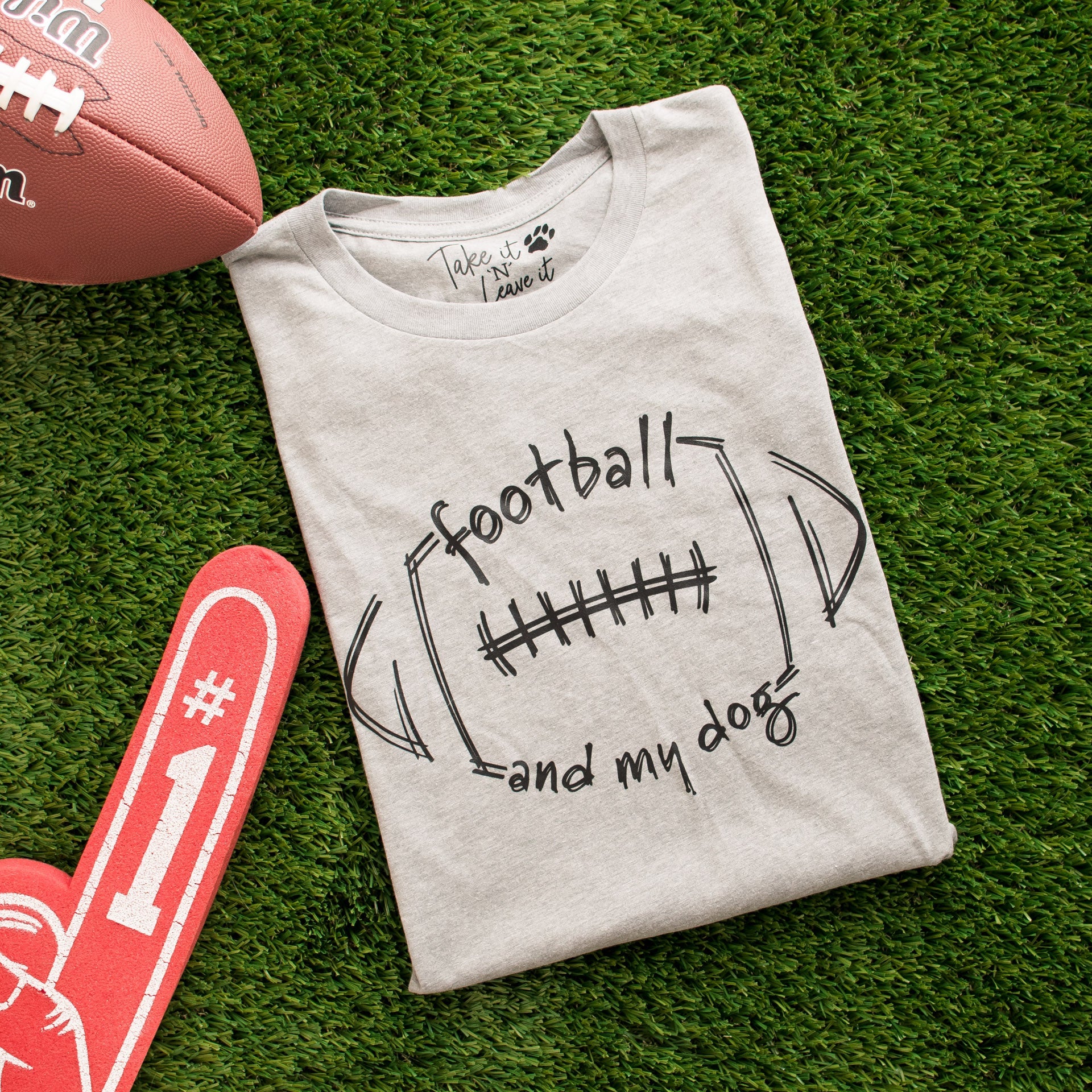 Folded t-shirt with 'football and my dog' text on grass with football and foam finger