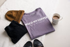 Purple t-shirt with humorous text, black pants, brown hat, and coffee cup on a white surface