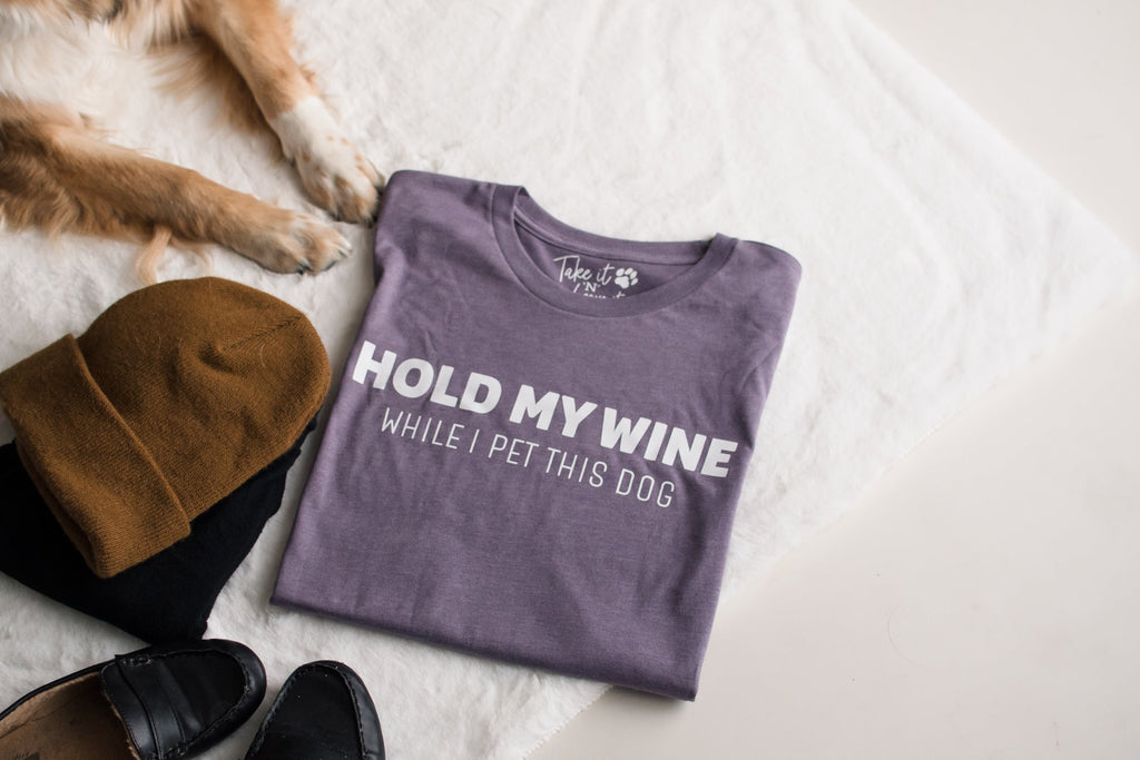 Folded t-shirt with hold my wine while I pet this dog text on a white surface next to a dog and slippers.