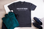 Black t-shirt with 'Hold my beer while I pet this cat' text, green jacket, and black shoes on a light wooden surface.