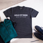 Black t-shirt with 'Hold my beer while I pet this cat' text, green jacket, and black shoes on a light wooden surface.