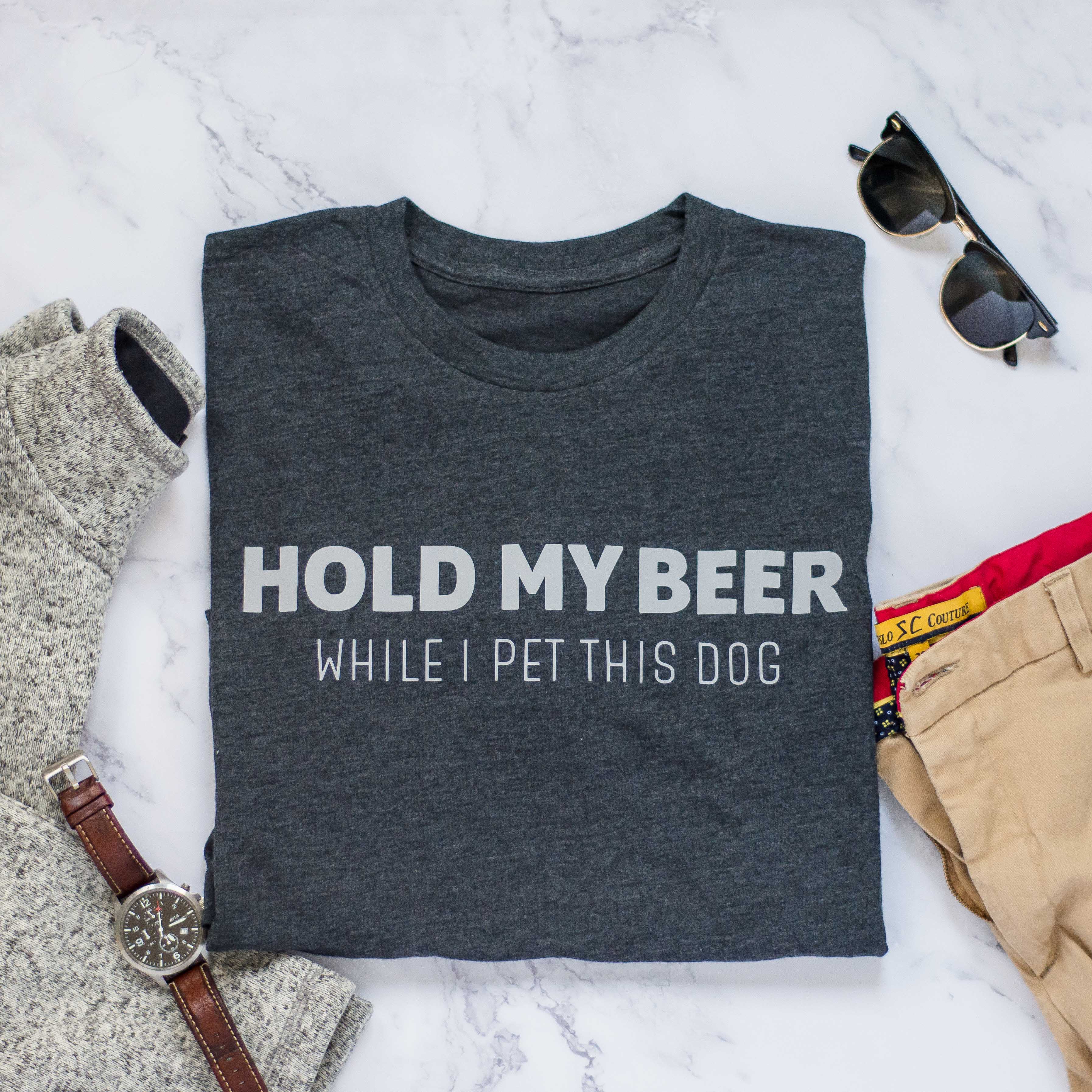 Dark gray t-shirt with text 'Hold my beer while I pet this dog' on a marble surface with accessories.