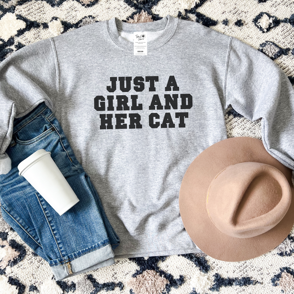 Gray sweatshirt with 'Just a Girl and Her Cat' text, paired with jeans, a hat, and a cup on a patterned surface.