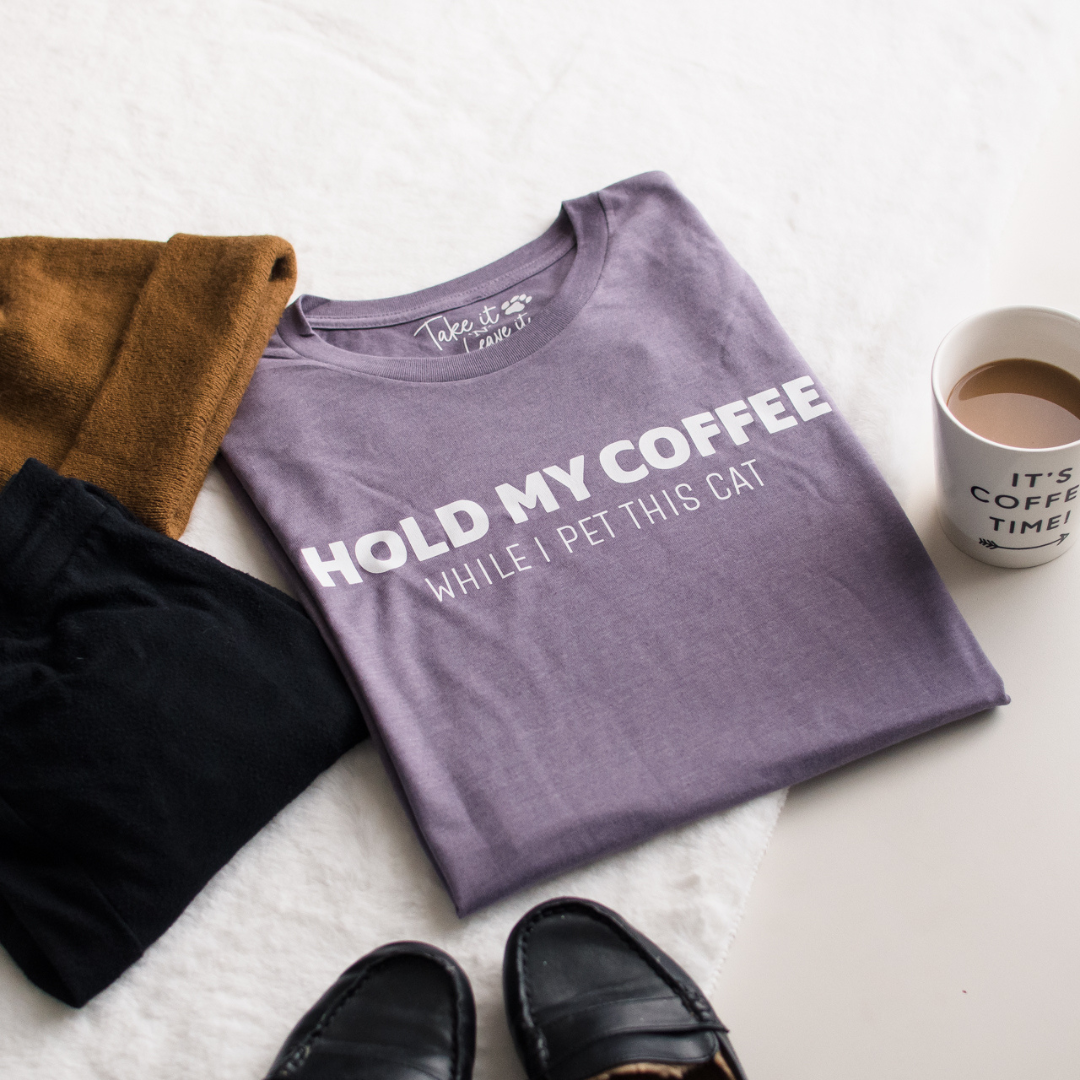 Folded purple t-shirt with text 'Hold my coffee while I pet this cat' on a white surface with coffee cup and shoes.