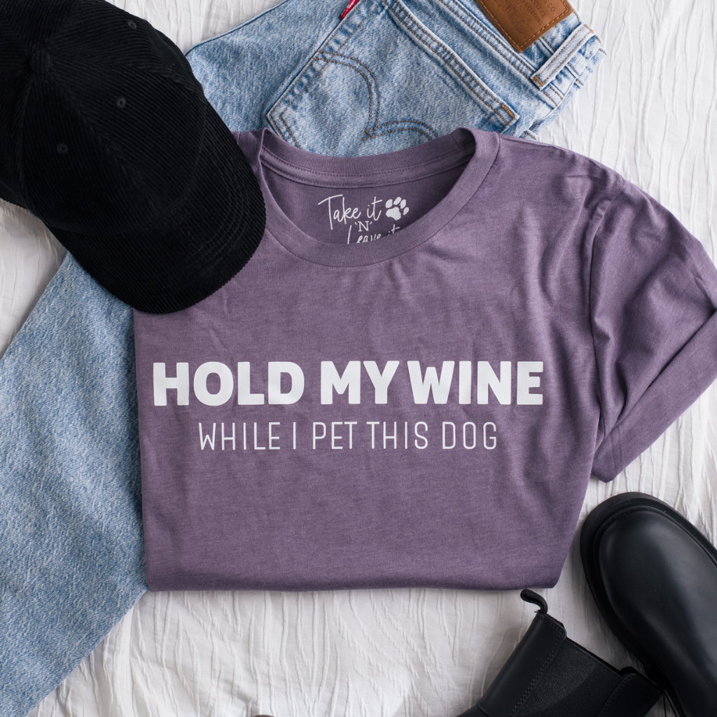 Purple t-shirt with 'Hold my wine while I pet this dog' text on a white surface with jeans and a cap.
