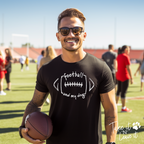 Man holding a football on a sports field wearing a black t-shirt with text.