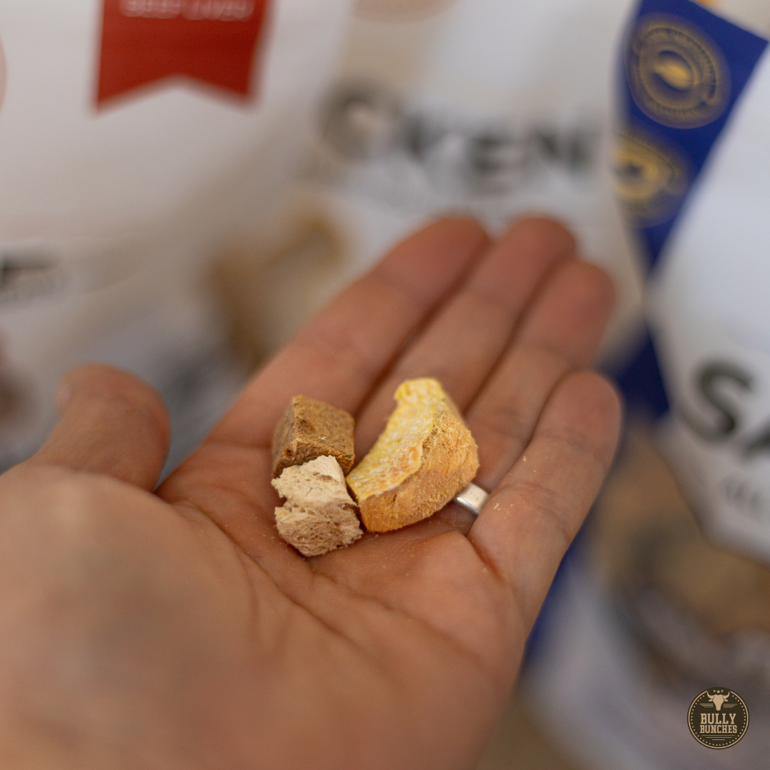 A close-up of a person's hand holding two small pieces of dog chews, with blurred dog food bags in the background.