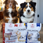 Two dogs sitting behind three bags of Bully Branded dog treats on a wooden floor in an indoor setting. The bags are labeled Beef, Chicken, and Salmon.