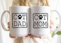 Two white mugs with 'Cat Dad' and 'Cat Mom' text held by a person.
