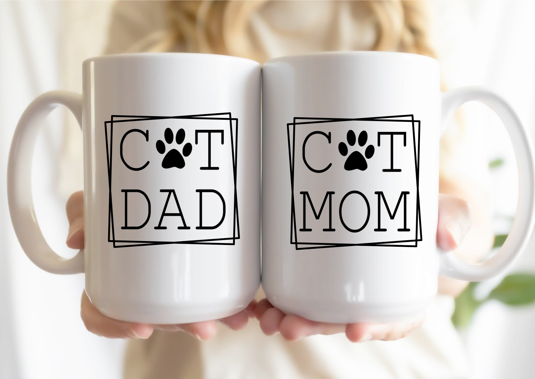 Two white mugs with 'Cat Dad' and 'Cat Mom' text held by a person.