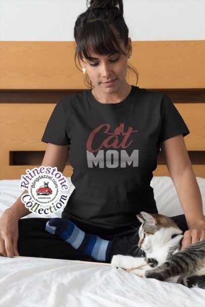 Woman wearing a 'Cat Mom' t-shirt sitting on a bed with a cat.