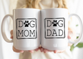 Two white mugs with 'Dog Mom' and 'Dog Dad' text held together.