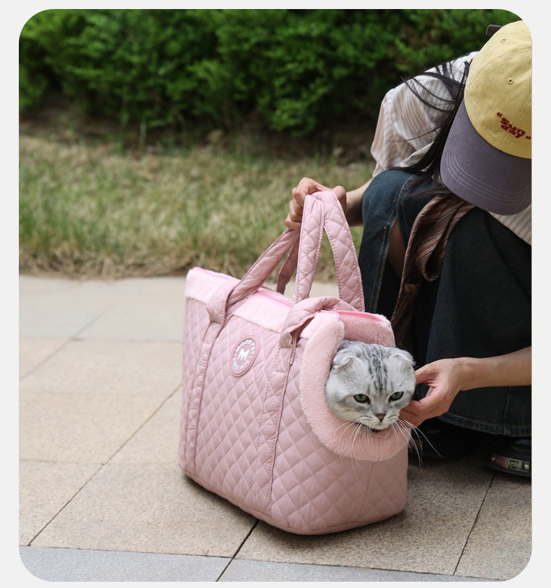 Person holding a pink quilted pet carrier with a cat peeking out, sitting on a pavement.