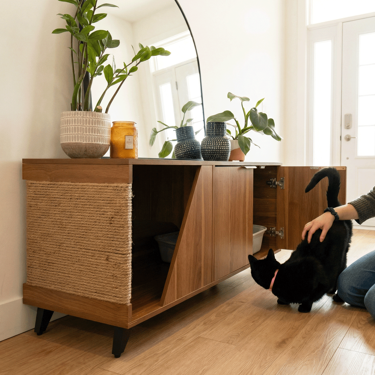Person interacting with a black cat next to a wooden cabinet with a cat tree inside