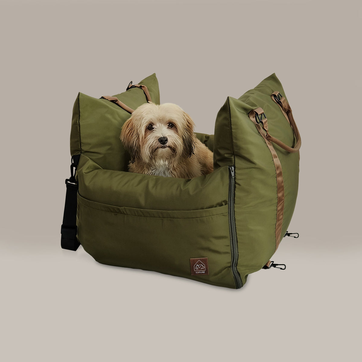 An olive green dog bed with brown handles and a small dog sitting inside, photographed against a solid light beige background.