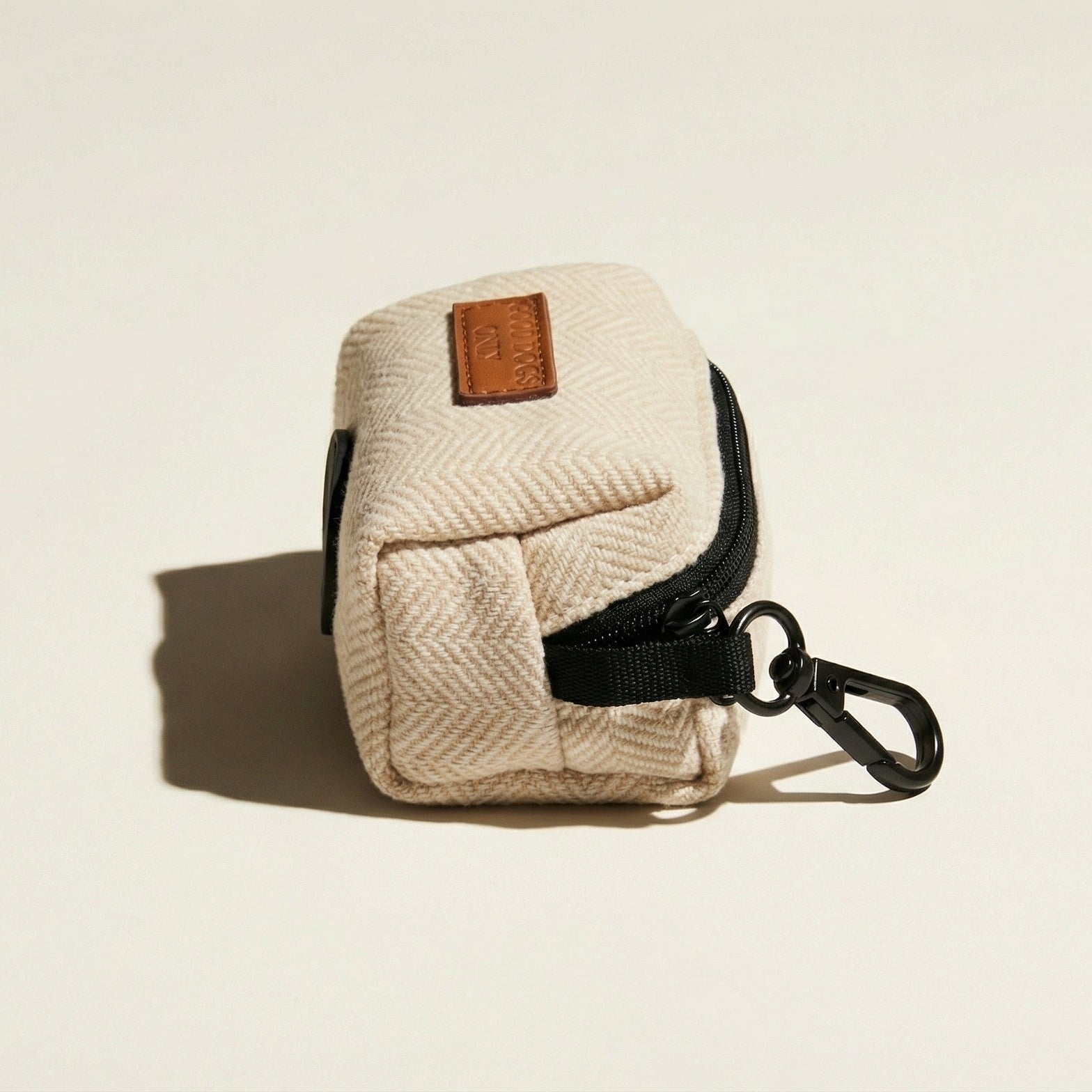 Beige fabric dog waste bag dispenser with black zipper and brown leather tag on a beige background