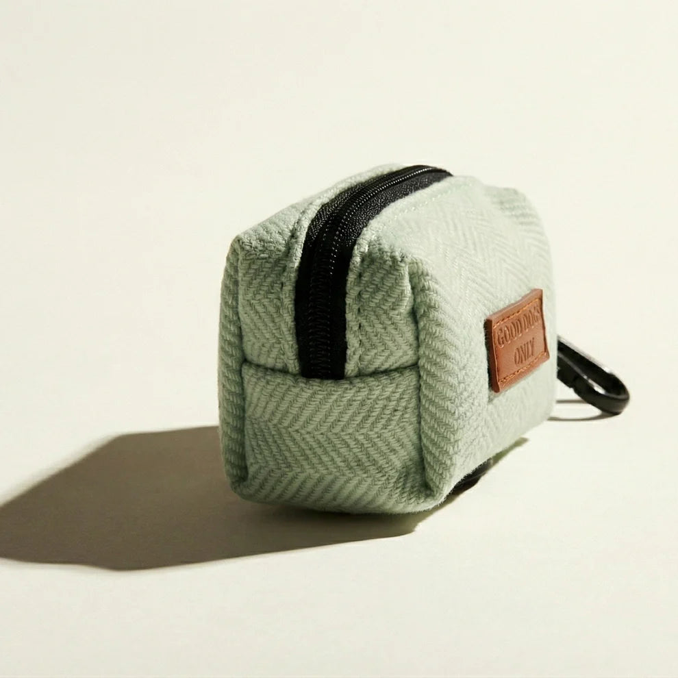 Green textured dog waste bag holder with a black zipper and brown leather patch on a beige background