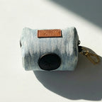 Denim dog waste bag holder with black bag and gold clip on a light gray background