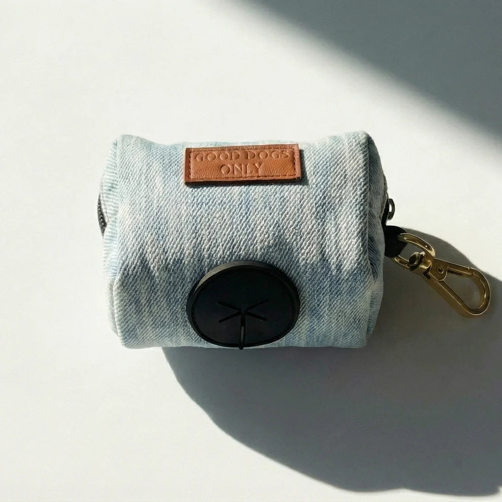 Denim dog waste bag holder with black bag and gold clip on a light gray background