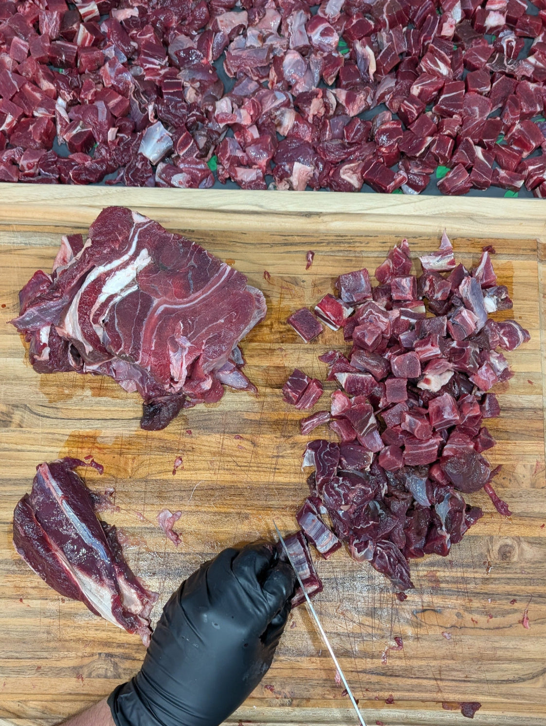Person cutting raw meat on a wooden cutting board with a tray of chopped meat in the background.