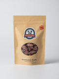 A stand-up kraft paper pouch of Dried & True Henational Hearts dog treats, featuring a circular window that displays the reddish-brown heart-shaped treats inside. The packaging has a blue and white logo in the center and a red circular seal on the upper right.