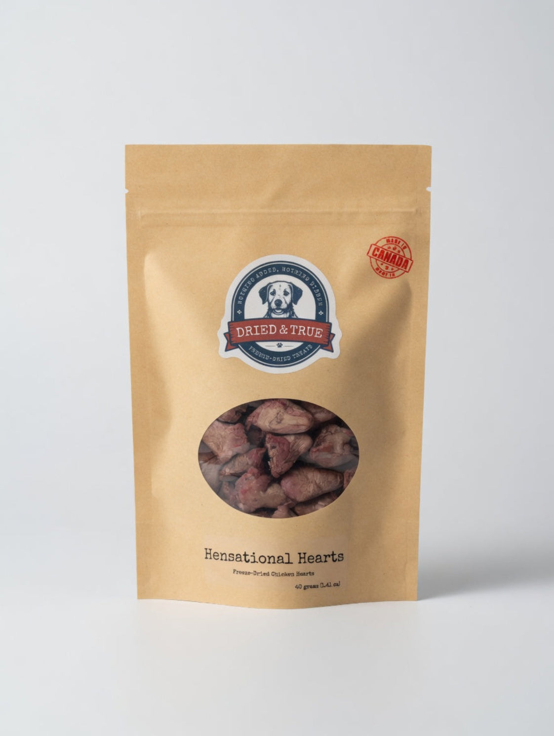 A stand-up kraft paper pouch of Dried & True Henational Hearts dog treats, featuring a circular window that displays the reddish-brown heart-shaped treats inside. The packaging has a blue and white logo in the center and a red circular seal on the upper right.