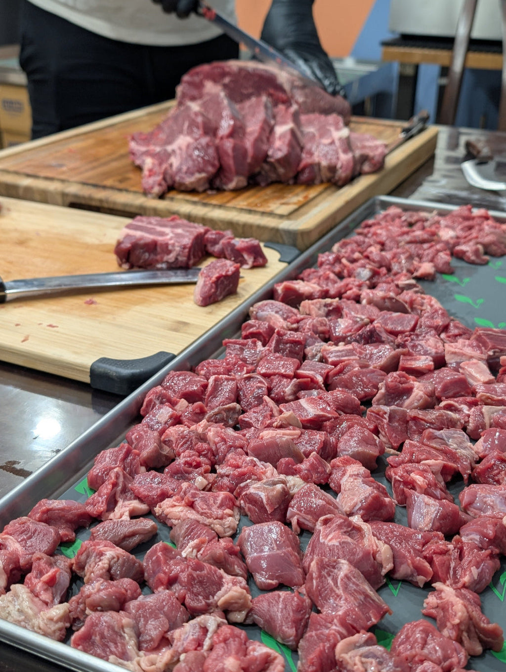 Raw meat being cut on a wooden cutting board with a tray of diced meat in the foreground.