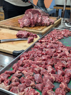 Raw meat being cut on a wooden cutting board with a tray of diced meat in the foreground.