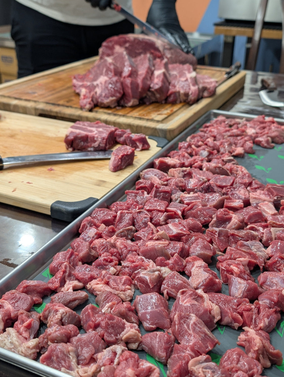 Raw meat being cut on a wooden cutting board with a tray of diced meat in the foreground.