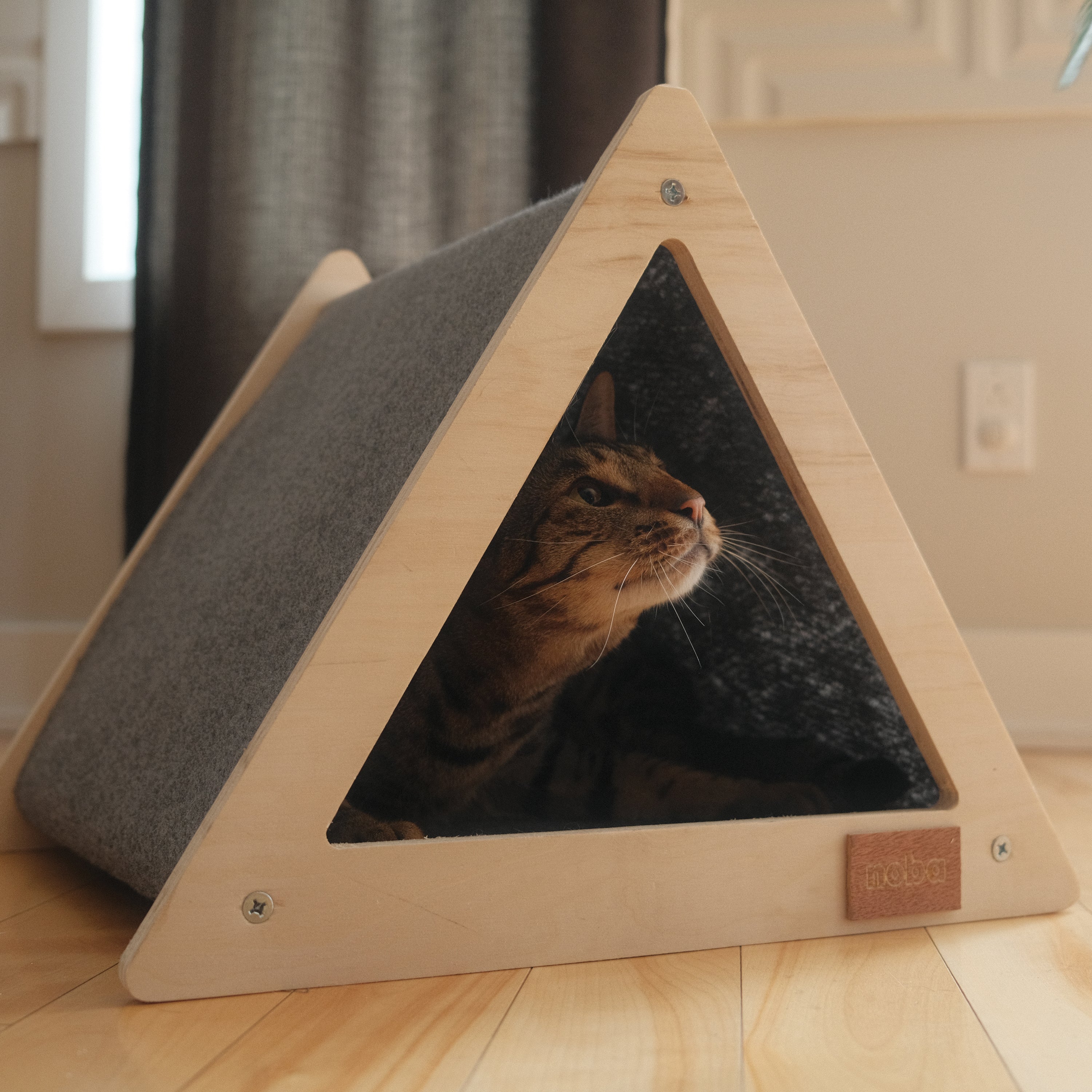 Triangular wooden pet bed with a cat peeking out on a wooden floor.