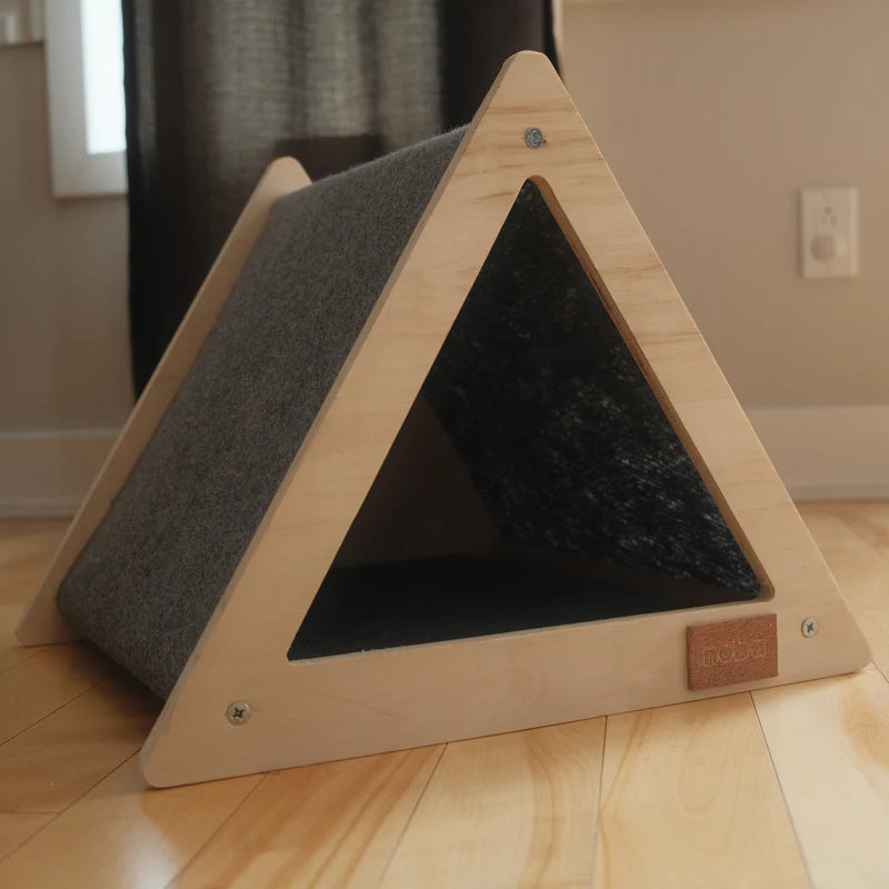 Triangular pet bed with wooden frame and gray cushion on a wooden floor.