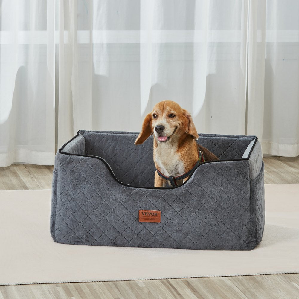 Dog sitting in a gray pet car seat with a visible brand logo, on a light-colored floor with curtains in the background.