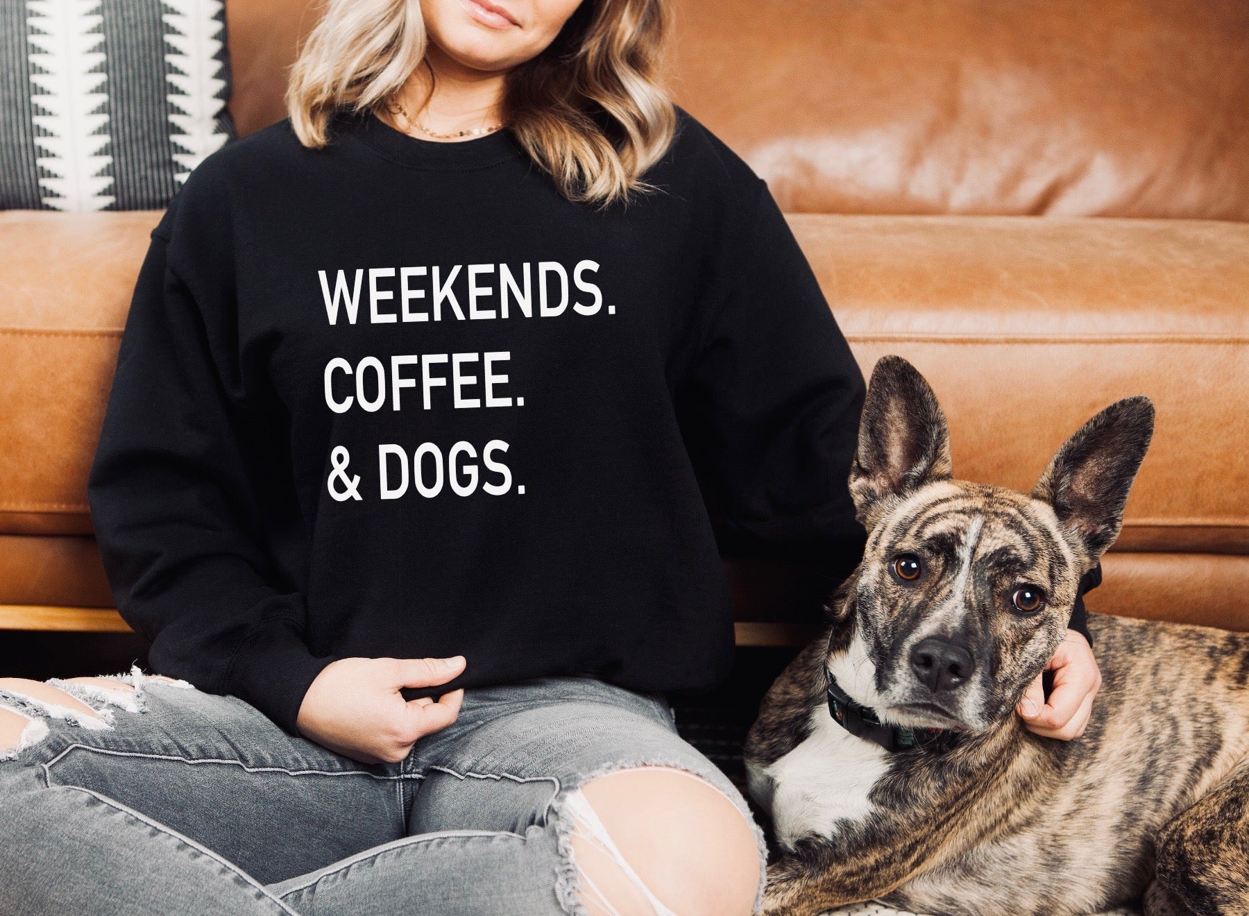 Person wearing a black sweatshirt with 'WEEKENDS. COFFEE. & DOGS.' text, sitting on a couch with a dog.